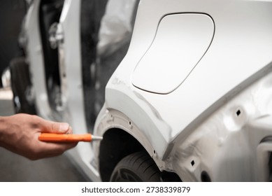 Close-up of a hand using an orange compressed air blowgun to remove dust from a white car's primed surface near the wheel arch inside an auto repair setting.