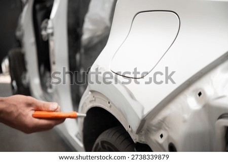 Close-up of a hand using an orange compressed air blowgun to remove dust from a white car's primed surface near the wheel arch inside an auto repair setting.