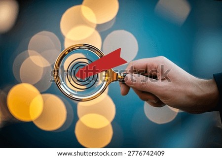 Close-up of a hand using a red arrow magnifying glass to highlight a golden target among scattered gold coins, symbolizing financial focus, investment strategy, and business success for marketing and advertising.