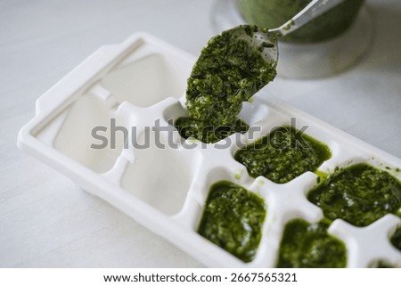 Close-up of hand using spoon to fill white ice cube tray with vibrant green pesto. Focus highlights the texture and color of sauce as it's prepared for freezing