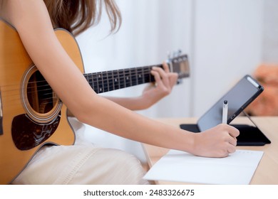 Closeup hand woman sitting on sofa looking tablet and compose a song for practice music with guitar in living room, woman watching lesson online about music and learning guitar, lifestyle concept. - Powered by Shutterstock