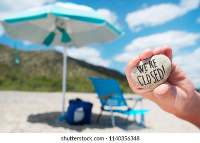 Closeup Of The Hand Of A Young Caucasian Man Holding A Stone With The Text We Are Closed Written In It, On The Beach, With An Umbrella And A Deck Chair In The Background