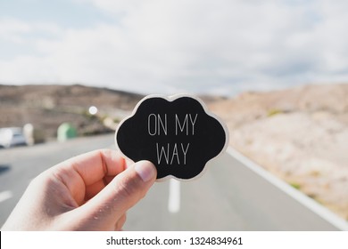 Closeup Of The Hand Of A Young Caucasian Man Holding A Black Signboard, In The Shape Of A Thought Bubble, With The Text On My Way Written In It, On A Road