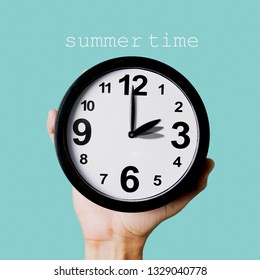 Closeup Of The Hand Of A Young Caucasian Man Holding A Clock, Which Is Being Adjusted One Hour, And The Text Summer Time Against A Blue Background