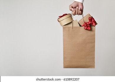 Closeup Of The Hand Of A Young Caucasian Man Carrying A Paper Shopping Bag Full Of Gift Boxes Ornamented With Red Ribbon Against An Off-white Background, And A Negative Space On The Left