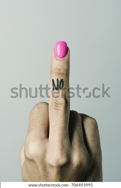 closeup of the hand of a young caucasian woman giving the middle finger with the word no handwritten in it, against an off-white background