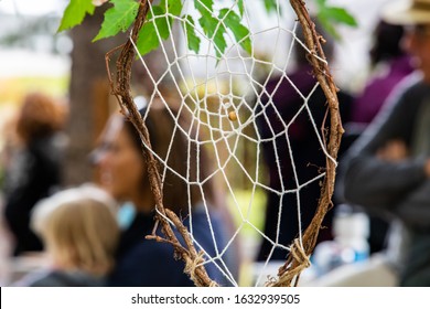 Closeup Of Handmade Dreamcatcher Hanging On Tree Branch With Defocus Group Of People Sitting And Looking At Something At World And Spoken Word Event