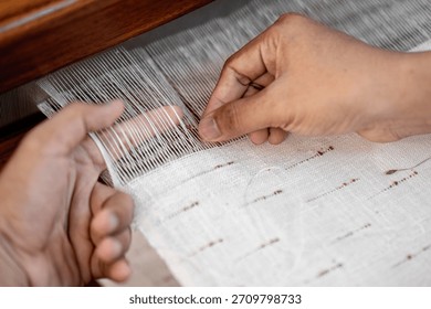 Close-up of hands adjusting fine threads on a traditional handloom during fabric weaving. Focus on manual technique and thread alignment.