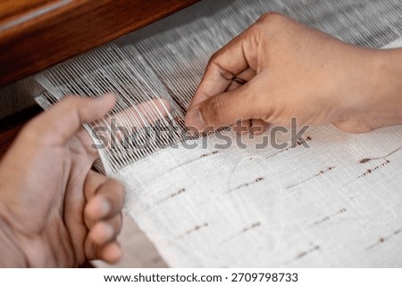 Close-up of hands adjusting fine threads on a traditional handloom during fabric weaving. Focus on manual technique and thread alignment.