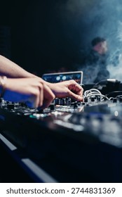 Close-up of a DJ’s hands adjusting knobs on a professional mixing console during a live performance in a nightclub. Blue stage lighting and atmospheric smoke create a dynamic nightlife scene, ideal for themes related to music, entertainment, events, and modern urban culture.
