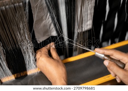Close-up of hands adjusting thin threads on a weaving loom using a guiding tool. The image shows detailed manual work in textile preparation.