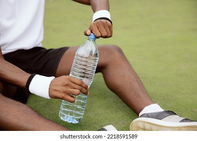 Close-up of the hands of an African sportsman holding a bottle of water and uncorking it. The guy is sitting on the grass of the tennis court and is wearing a white T-shirt, wristbands, and black shorts.
