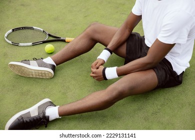 Close-up of the hands of African-looking tennis player sitting on the grass of tennis court. Next to him lies tennis racket and ball. Guy is wearing white T-shirt, black shorts and sneakers.