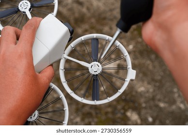 Close-up of hands assembling a compact white drone with black propellers using a screwdriver on an outdoor textured ground surface focusing on mechanical assembly process