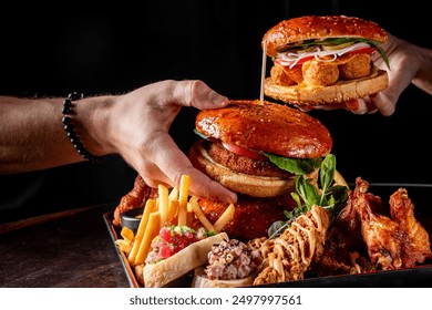 Close-up of hands assembling a large, multi-layered burger with various fillings. The tray also includes sides like fries and chicken wings. Perfect for food and culinary themes.