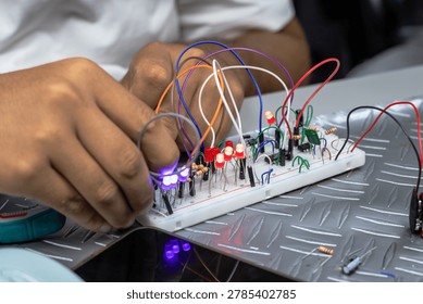 Close-up of hands assembling and testing an electronic circuit on a breadboard with glowing LEDs, jumper wires, and components in a workshop or electronics lab.