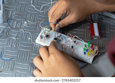 Close-up of hands assembling and testing an electronics circuit on a breadboard with wires, LEDs, and battery on a metal surface. Educational STEM project concept.