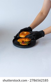Close-up of hands in black gloves assembling a gourmet bagel sandwich, placing the top bun over a crispy fried chicken fillet drizzled with creamy spicy sauce and red cabbage.