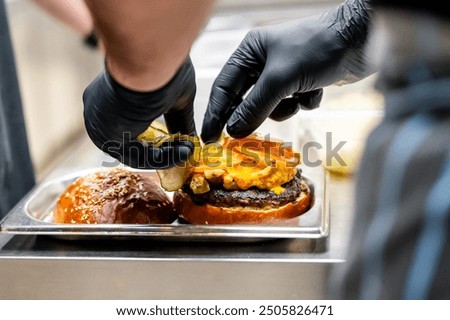 Close-up of hands in black gloves assembling a burger with cheese and a sesame bun on a metal tray, highlighting fresh ingredients. Perfect for food preparation, culinary arts, and restaurant themes.