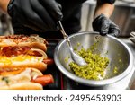 Close-up of hands in black gloves preparing hot dogs with bacon and cheese toppings, adding relish. Street food preparation, culinary concept. Perfect for food blogs, culinary websites