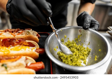 Close-up of hands in black gloves preparing hot dogs with bacon and cheese toppings, adding relish. Street food preparation, culinary concept. Perfect for food blogs, culinary websites - Powered by Shutterstock