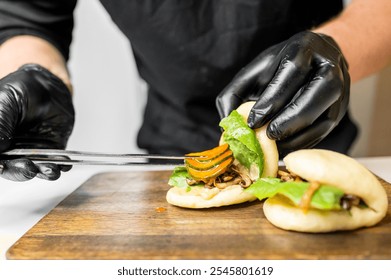 Close-up of hands in black gloves preparing a gourmet sandwich with fresh ingredients, including lettuce and sliced vegetables, on a wooden cutting board. Food preparation, culinary arts - Powered by Shutterstock