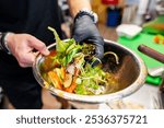 A close-up of hands in black gloves tossing a fresh salad with mixed greens and sliced tomatoes in a metal bowl, highlighting healthy eating.