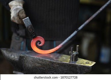 A close-up of the hands of a Blacksmith forging a curl from a red-hot flattened blank with a hammer. Handmade in the forge concept