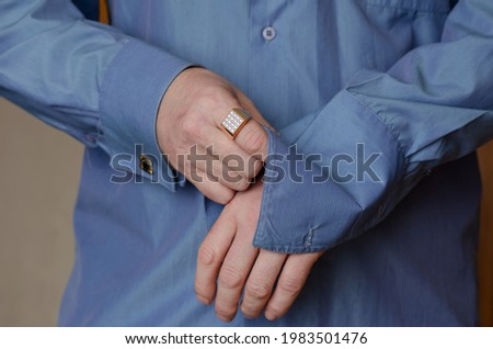 Close-up of hands buttoning a cufflink on the sleeve of a blue shirt. A middle-aged adult man with a gold ring on his index finger. Person holding hands in middle section of body. Lifestyle. Indoors.