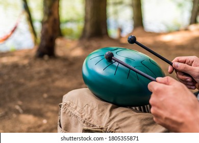 Close-up with the hands of a Caucasian musician holding drum sticks and playing a modern hand pan steel tongue drum percussion instrument in the middle of nature.