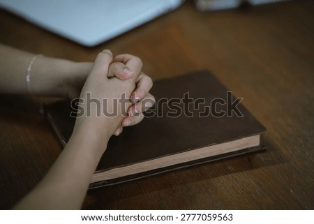 Close-up of hands clasped in prayer over a closed book on a wooden table, symbolizing faith, reflection, and spiritual devotion in a quiet indoor setting.