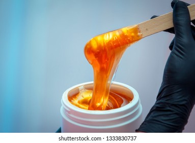 Close-up hands of cosmetologist in black gloves preparing golden colored waxing paste on spatula for sugaring depilation, hair removal beauty procedure.