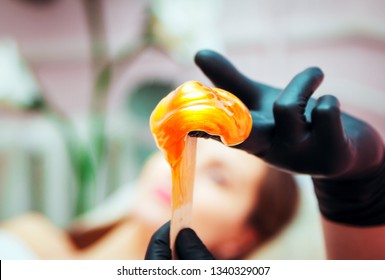 Close-up hands of cosmetologist in black gloves preparing golden colored waxing paste on spatula for sugaring depilation, hair removal beauty procedure.