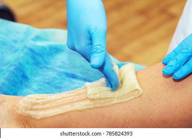Close-up hands of cosmetologist in blue gloves applying paste for sugaring depilation, hair removal beauty procedure.