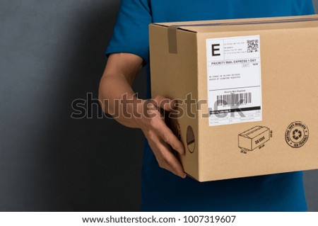 Closeup hands of delivery man holding package to deliver. Courier hand holding brown box isolated on grey background. Detail of delivery man carrying cardboard parcel with label with copy space.
