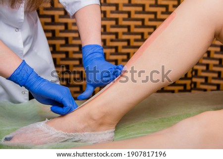 Close-up of the hands of a depilation master in blue gloves remove hair with a paper strip with wax on the client's legs. Hair removal process on legs in a beauty salon.