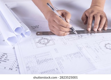 Close-up of hands drafting technical plans with a pen and ruler on engineering drawings and rolled blueprints at a desk.