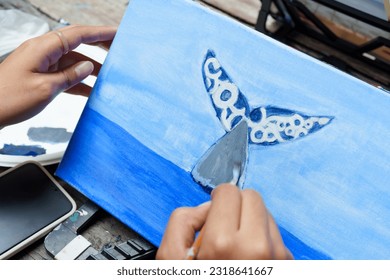 Close-up. Hands of female apprentice painter holding paintbrush. Paint  whales and sea on four corners of wooden cloth. Decorate colors beautifully before sending them to teacher. in school art class