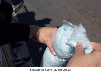 Close-up of hands gently pulling light blue cotton candy from a plastic bag outdoors. Bright natural daylight highlights the fluffy texture and vibrant color. This image conveys a sweet, joyful, nostalgic treat atmosphere.