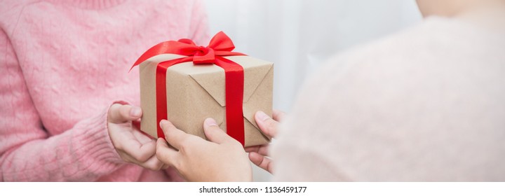 Closeup Hands Giving Surprise Gift Box, Two Woman Delivery Gift Brown Package Box With Red Ribbon. Boxing Day Holiday Birthday Christmas Concept Panoramic Banner