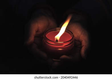 Close-up Of Hands Holding A Burning Candle In The Dark. Adult Woman With A Red Candle Against A Black Background. Candle In A Glass Jar. Selective Focus.