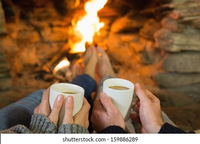 Close-up of hands holding coffee cups in front of lit fireplace