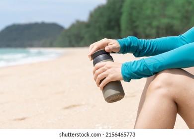 Closeup of woman’s hands holding insulated stainless reusable water bottle sitting in on beach on sunny day. Bring your own bottle, Eco friendly, zero waste and green living lifestyle. - Shutterstock ID 2156465773