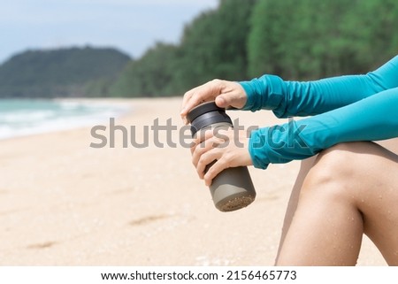 Closeup of woman’s hands holding insulated stainless reusable water bottle sitting in on beach on sunny day. Bring your own bottle, Eco friendly, zero waste and green living lifestyle.