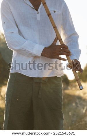 Close-up of a man’s hands holding and playing a ney flute, surrounded by the peaceful atmosphere of an olive field, sunlight glowing in the background