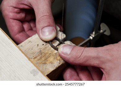 Close-up of hands holding a ring in place on a wooden block while using a fine saw blade for precision cutting. The hands are steady, carefully working with the metal piece in a jewelry-making setting - Powered by Shutterstock