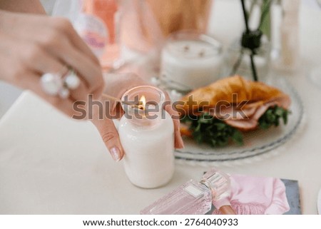 Close-up of hands lighting a white aromatic candle with a long match. A glass perfume bottle, greeting card, and croissant sandwich complete the elegant festive table setting.