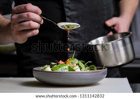 Close-up of the hands of a male chef on a black background. Pour sauce from the spoon on the salad dish. Food decoration.