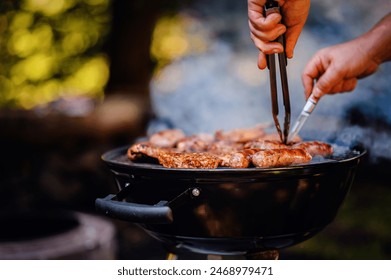 Close-up of hands masterfully flipping meats on a smoky grill, set against a blurred natural backdrop - Powered by Shutterstock