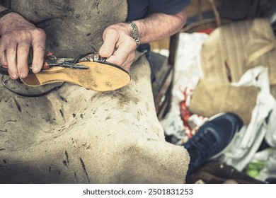 Close-up of the hands of an old master shoemaker in a leather apron using pliers to repair a shoe. In his messy workshop. Concept: Shoe repair circular economy and ecology against planned obsolescence - Powered by Shutterstock